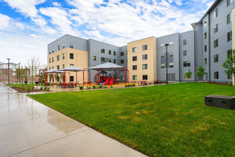 Modern multi-story apartment complex with gray and beige exterior siding, featuring a landscaped courtyard with green lawn, covered picnic areas, playground equipment, and concrete walkways under a partly cloudy sky.